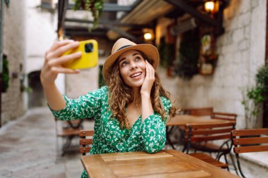 Genç bir kadın elinde dijital kamerayla yazlık kafede otururken selfie çekiyor. Moda, güzellik, blog, turizm.