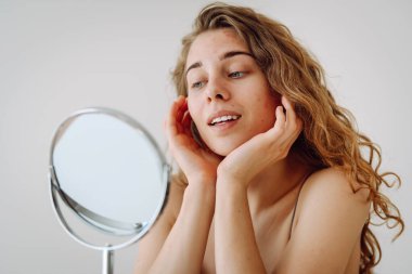 Güzel bir kadın aynaya bakıyor, elleriyle yüzüne dokunuyor, sivilceleri ve pigmentasyonu inceliyor. Rosacea ve couperose konsepti. Kozmetik bakım. Alerjik reaksiyon.
