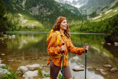 Sarı sırt çantası ve yürüyüş çubuklarıyla dağ gölünün kıyısına doğru yürüyen bir kadın gezgin. Dağ manzarasının tadını çıkarıyor. Aktif yaşam tarzı. Seyahat tutkusu.