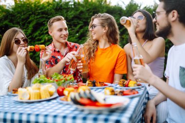 Bir grup mutlu genç arkadaş dışarıda piknik yapıyor. İnsanlar barda ızgara yaparken eğleniyor ve kutluyorlar. Tatil, piknik, hafta sonu, doğa