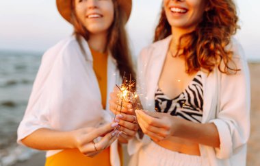 Kumsaldaki genç bayanın ellerindeki maytaplar. Kumsalda kutlama yapan iki kadın. Yaz tatili, parti, tatil, rahatlama ve yaşam tarzı konsepti.