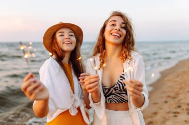 Kumsaldaki genç bayanın ellerindeki maytaplar. Kumsalda kutlama yapan iki kadın. Yaz tatili, parti, tatil, rahatlama ve yaşam tarzı konsepti.