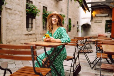 Akıllı telefon tutan ve kahve dükkanında cep telefonundan mesaj okuyan mutlu genç bir kadın. İnsanlar, moda, yaşam tarzı, seyahat ve tatil kavramı.