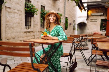 Akıllı telefon tutan ve kahve dükkanında cep telefonundan mesaj okuyan mutlu genç bir kadın. İnsanlar, moda, yaşam tarzı, seyahat ve tatil kavramı.