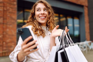 Genç kadın cep telefonu kullanıyor ve alışveriş çantasıyla bekliyor. Tüketim, satış, kara cuma. İnternetten alışveriş.