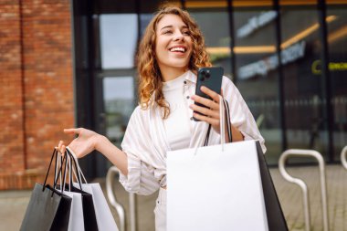 Genç kadın cep telefonu kullanıyor ve alışveriş çantasıyla bekliyor. Tüketim, satış, kara cuma. İnternetten alışveriş.