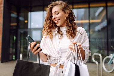 Genç kadın cep telefonu kullanıyor ve alışveriş çantasıyla bekliyor. Tüketim, satış, kara cuma. İnternetten alışveriş.