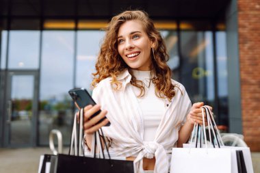 Genç kadın cep telefonu kullanıyor ve alışveriş çantasıyla bekliyor. Tüketim, satış, kara cuma. İnternetten alışveriş.