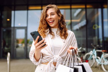 Genç kadın cep telefonu kullanıyor ve alışveriş çantasıyla bekliyor. Tüketim, satış, kara cuma. İnternetten alışveriş.