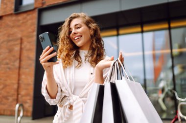 Genç kadın cep telefonu kullanıyor ve alışveriş çantasıyla bekliyor. Tüketim, satış, kara cuma. İnternetten alışveriş.