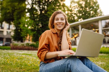 Genç bir kadın parkta oturmuş video görüşmesiyle iletişim kuruyor. Laptop bilgisayarına bakan bir kız, online konferans mesafesinde ofis sohbeti, sanal eğitim. Serbest çalışma..