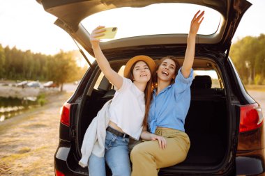 Genç bir kadın arabanın bagajında oturur ve selfie çeker. Araba yolculuğunda şık modeller. Teknoloji, blog, seyahat kavramı.