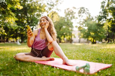 Parkın doğasında kadın sporcu. Yoga minderinde dinleniyor. Cep telefonu kullanıyor, uygulama egzersizi yapıyor. Çevrimiçi sohbet. Etkin yaşam biçimi.