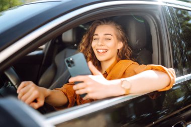Sürücü koltuğunda oturan ve akıllı telefon kullanan güzel bir genç kadın. Araba paylaşımı, kiralama servisi ya da taksi uygulaması. Yaşam tarzı, seyahat, turizm, aktif yaşam.