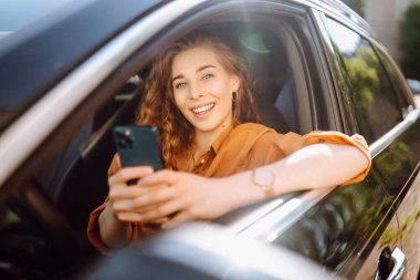 Sürücü koltuğunda oturan ve akıllı telefon kullanan güzel bir genç kadın. Araba paylaşımı, kiralama servisi ya da taksi uygulaması. Yaşam tarzı, seyahat, turizm, aktif yaşam.