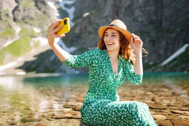 Cep telefonlu bir kadın gezgin gölün arka planında selfie çekiyor. Yaz tatilinde turist. Macera, tatil.