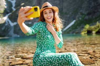 Cep telefonlu bir kadın gezgin gölün arka planında selfie çekiyor. Yaz tatilinde turist. Macera, tatil.