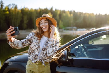 Genç bayan turist otomobille selfie çekiyor. Araba gezisinde fotoğraf çekmek için mola veriyoruz. Aktif yaşam tarzı, blog yazma, doğa, hafta sonu.