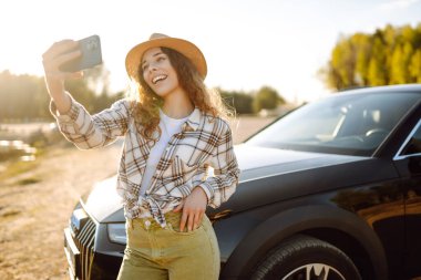 Genç bayan turist otomobille selfie çekiyor. Araba gezisinde fotoğraf çekmek için mola veriyoruz. Aktif yaşam tarzı, blog yazma, doğa, hafta sonu.