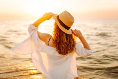 Gün batımında sahilde yürüyen mutlu bir kadın. Gün batımında deniz manzarası ve güzel bir gökyüzü. Seyahat, hafta sonu, rahatlama ve yaşam tarzı konsepti.