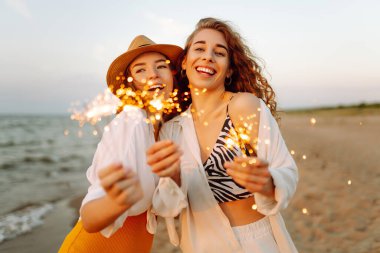 Kumsaldaki genç kadınların ellerindeki maytaplar. Kumsalda havai fişeklerle yürüyen mutlu kadınlar. Yaz tatili, parti, tatil, rahatlama ve yaşam tarzı konsepti.