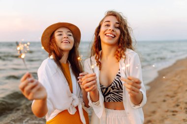 Kumsaldaki genç kadınların ellerindeki maytaplar. Kumsalda havai fişeklerle yürüyen mutlu kadınlar. Yaz tatili, parti, tatil, rahatlama ve yaşam tarzı konsepti.