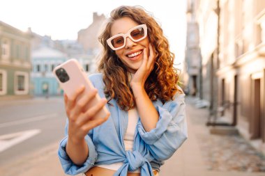 Yaşlı caddede yürürken gülümseyen ve akıllı telefon kullanan genç bir kadın. İletişim, sosyal ağlar, blog yazma, online alışveriş kavramı.