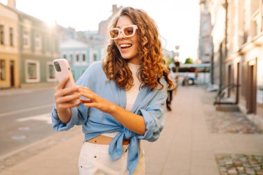 Yaşlı caddede yürürken gülümseyen ve akıllı telefon kullanan genç bir kadın. İletişim, sosyal ağlar, blog yazma, online alışveriş kavramı.
