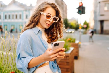 Yaşlı caddede yürürken gülümseyen ve akıllı telefon kullanan genç bir kadın. İletişim, sosyal ağlar, blog yazma, online alışveriş kavramı.