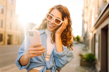 Yaşlı caddede yürürken gülümseyen ve akıllı telefon kullanan genç bir kadın. İletişim, sosyal ağlar, blog yazma, online alışveriş kavramı.