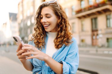 Yaşlı caddede yürürken gülümseyen ve akıllı telefon kullanan genç bir kadın. İletişim, sosyal ağlar, blog yazma, online alışveriş kavramı.