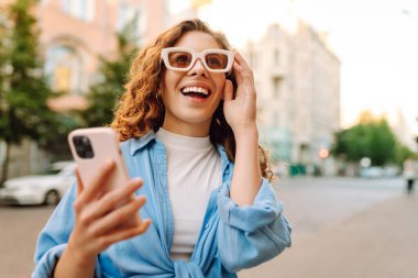 Yaşlı caddede yürürken gülümseyen ve akıllı telefon kullanan genç bir kadın. İletişim, sosyal ağlar, blog yazma, online alışveriş kavramı.