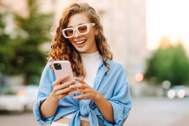 Yaşlı caddede yürürken gülümseyen ve akıllı telefon kullanan genç bir kadın. İletişim, sosyal ağlar, blog yazma, online alışveriş kavramı.