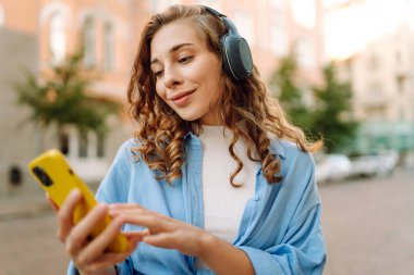 Kablosuz kulaklıklı mutlu, rahat bir kadın şehrin güneşli sokaklarında en sevdiği müziği seçiyor. Seyahat, turizm, teknoloji, blog konsepti.
