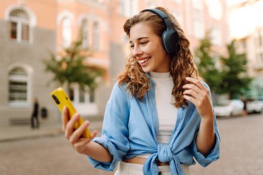 Kablosuz kulaklıklı mutlu, rahat bir kadın şehrin güneşli sokaklarında en sevdiği müziği seçiyor. Seyahat, turizm, teknoloji, blog konsepti.