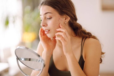 Derisi sorunlu genç bir kadın aynaya bakıyor. Sivilce terapisi kavramı, yara izleri, yüzünde iltihap, sorunlu cilt. Güzellik bakımı.