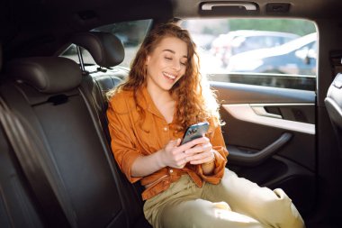 Genç bayan arka yolcu koltuğunda oturuyor ve akıllı telefonuyla konuşuyor. Teknoloji kavramı, araba ve iş seyahati..