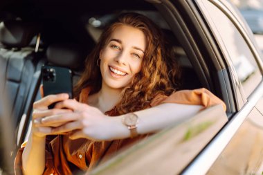 Genç bayan arka yolcu koltuğunda oturuyor ve akıllı telefonuyla konuşuyor. Teknoloji kavramı, araba ve iş seyahati..