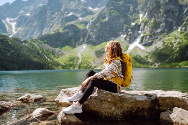 Büyük sarı sırt çantalı dişi turist güneşli havada dağ gölü manzarasının tadını çıkarıyor. Yürüyüş, seyahat, tatil konsepti.