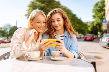 İki genç kadın, kız kardeş, en iyi arkadaşlar akıllı telefondan haber sosyal medya fotoğrafları gösteriyorlar kafede oturuyor ve kahvaltı sırasında kahve içiyorlar. Moda, güzellik, blog, turizm.