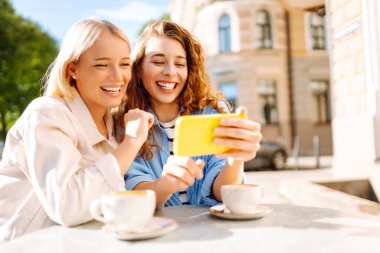 İki genç kadın, kız kardeş, en iyi arkadaşlar akıllı telefondan haber sosyal medya fotoğrafları gösteriyorlar kafede oturuyor ve kahvaltı sırasında kahve içiyorlar. Moda, güzellik, blog, turizm.