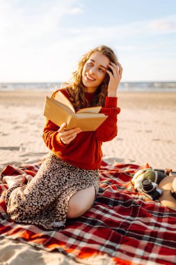 Kitap okuyan romantik bir kadın ekose üzerinde oturur, deniz kumsalı arka planında piknik yapar. İnsanlar, tatil, yaşam tarzı, yolculuk konsepti.