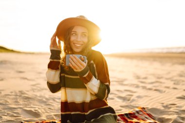 Genç mutlu turist plajda piknikte oturup sıcak bir şeyler içer. İnsanlar, tatil, yaşam tarzı, yolculuk konsepti.