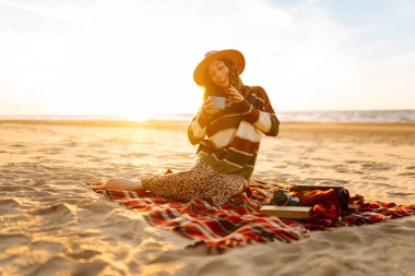 Genç mutlu turist plajda piknikte oturup sıcak bir şeyler içer. İnsanlar, tatil, yaşam tarzı, yolculuk konsepti.