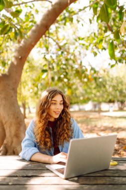Genç bir kadın dizüstü bilgisayarla parkta oturuyor. Uzaktan çalışma ya da eğitim. Serbest, çevrimiçi kurs.