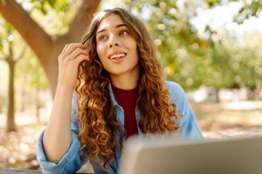 Genç bir kadın dizüstü bilgisayarla parkta oturuyor. Uzaktan çalışma ya da eğitim. Serbest, çevrimiçi kurs.