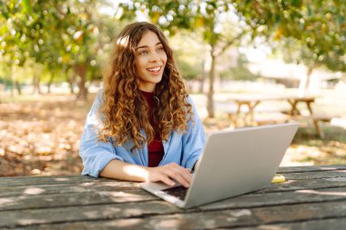 Genç bir kadın dizüstü bilgisayarla parkta oturuyor. Uzaktan çalışma ya da eğitim. Serbest, çevrimiçi kurs.