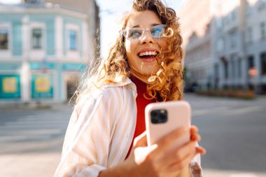 Gülümseyen genç bir kadın eski caddede yürüyor, selfie çekiyor, arkadaşlarla iletişim kuruyor. Ağlar, bloglar, online alışveriş kavramı.