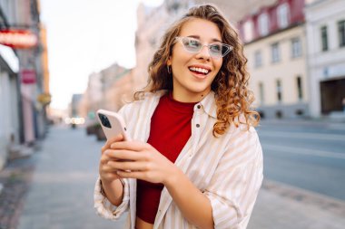 Gülümseyen genç bir kadın eski caddede yürüyor, selfie çekiyor, arkadaşlarla iletişim kuruyor. Ağlar, bloglar, online alışveriş kavramı.