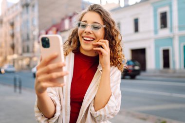 Gülümseyen genç bir kadın eski caddede yürüyor, selfie çekiyor, arkadaşlarla iletişim kuruyor. Ağlar, bloglar, online alışveriş kavramı.